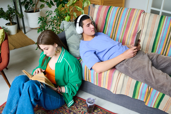 A zoomer girl in multi colored clothes is reading a book sitting on the carpet near the sofa on which a young man in white headphones is lying with a tablet in his hands